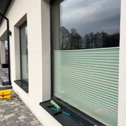 PARIX - Umyte okno z żaluzjami w nowoczesnym domu. Na parapecie leżą narzędzia do mycia okien: ściągaczka, myjka. Widoczna kostka brukowa i żółty pojemnik.