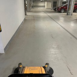 PARIX - Żółta zamiatarka przemysłowa w trakcie czyszczenia posadzki w garażu podziemnym. Widoczne ślady po czyszczeniu, w tle zaparkowane samochody.