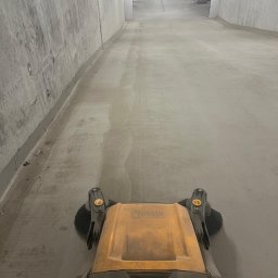 PARIX - Maszyna czyszcząca Ulsonix w trakcie pracy na betonowej posadzce w garażu podziemnym. Widoczne ślady po czyszczeniu, surowe betonowe ściany i oświetlenie.