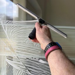 PARIX - Mycie okna z bliska: dłoń z raklem usuwa pianę z szyby, widoczne dwa zegarki na nadgarstku, okno z pionowymi żaluzjami w tle, etap czyszczenia.