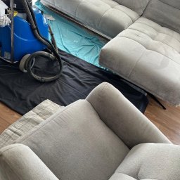 PARIX - Profesjonalne urządzenie do czyszczenia tapicerki obok jasnoszarego narożnika i fotela, zabezpieczone folią i czarną matą. Widok z góry na sprzęt i meble.