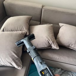 PARIX - Szczegółowe ujęcie dyszy odkurzacza piorącego na tle szarej, welurowej kanapy z poduszkami. Widoczne kontrolki urządzenia i fragment dłoni operatora.