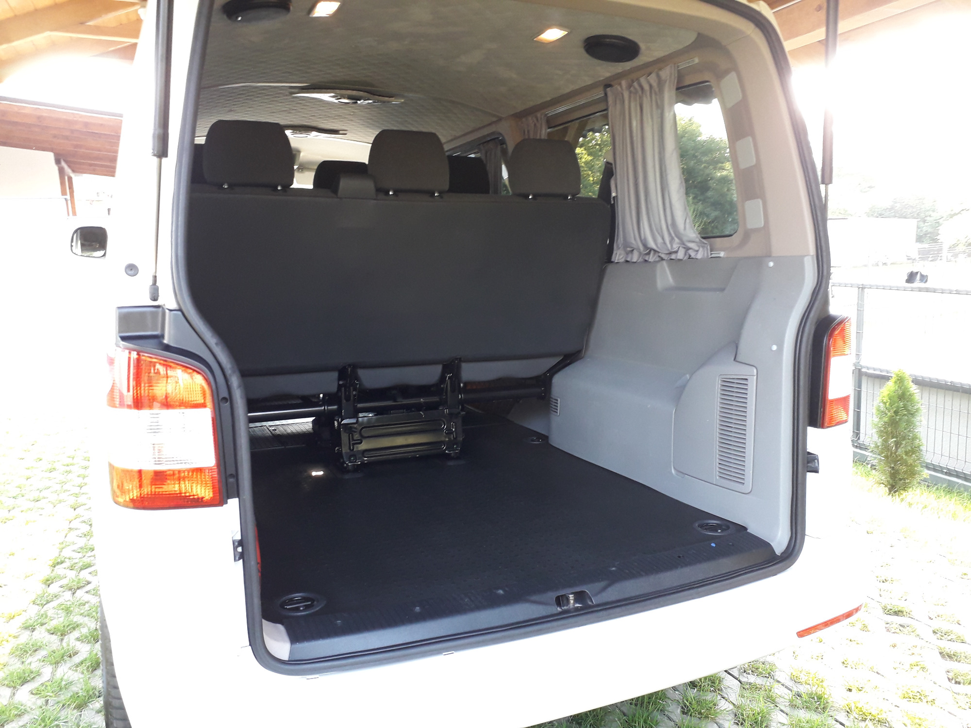 Wnętrze białego busa z rozłożonymi siedzeniami, widoczne szare zasłonki w oknach i czarna podłoga, światło słoneczne wpadające do środka.