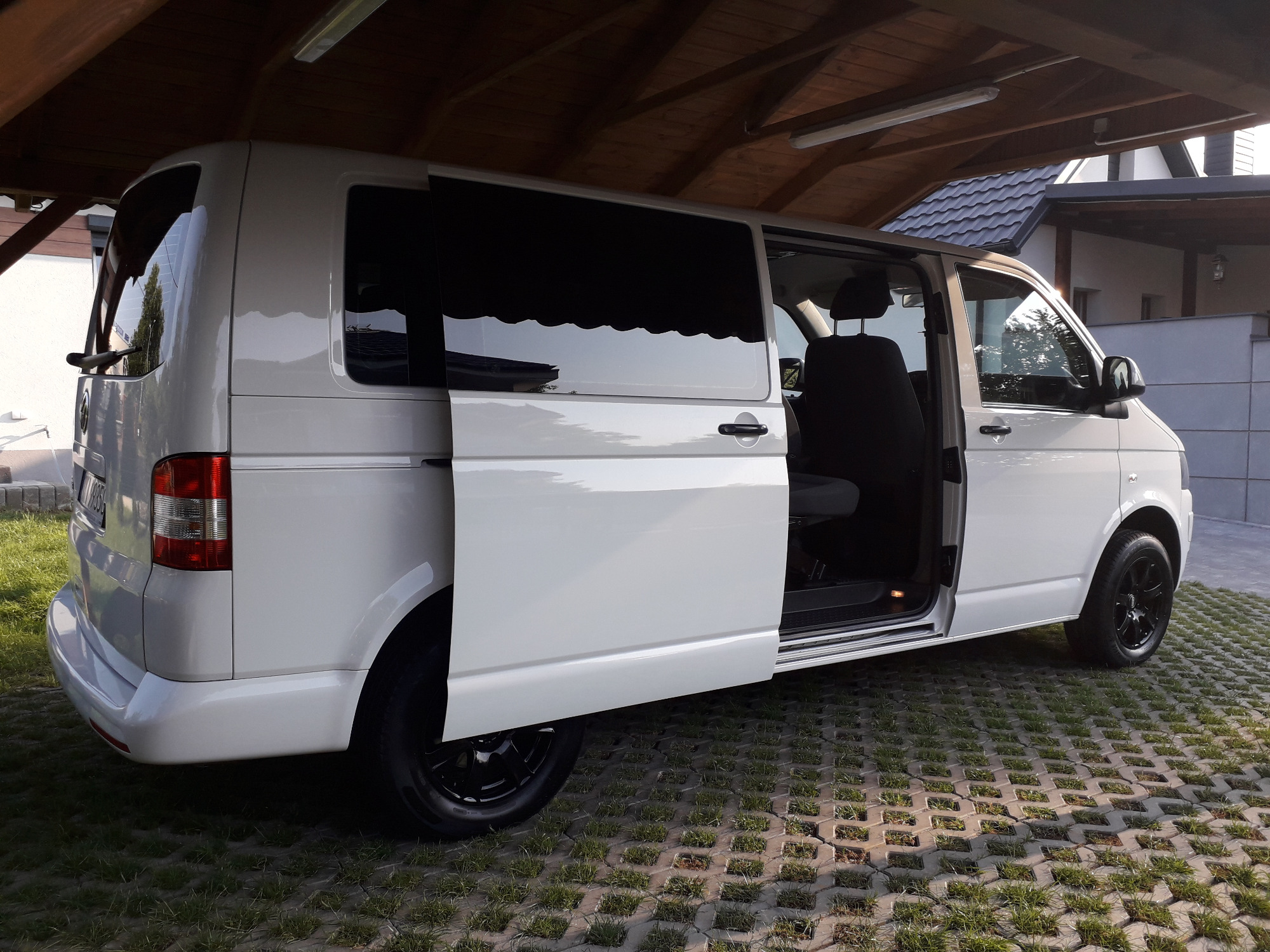 Biały van z otwartymi przesuwnymi drzwiami, zaparkowany na ażurowych płytach trawnikowych pod drewnianą wiatą.