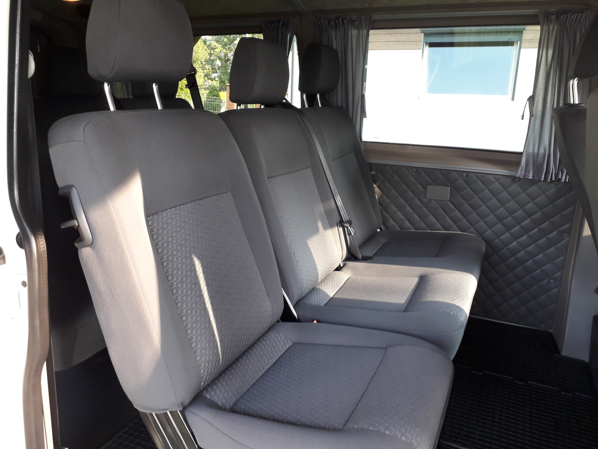 Wnętrze busa z trzema rzędami szarych foteli z zagłówkami i pasami bezpieczeństwa, widok z boku na tylne siedzenia, okno z zasłoną i pikowana tapicerka boczna.