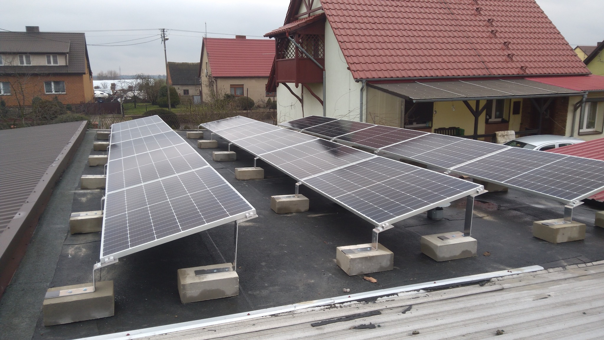 Panele słoneczne zamontowane na płaskim dachu z betonowymi blokami obciążającymi, w tle zabudowa jednorodzinna w pochmurny dzień.