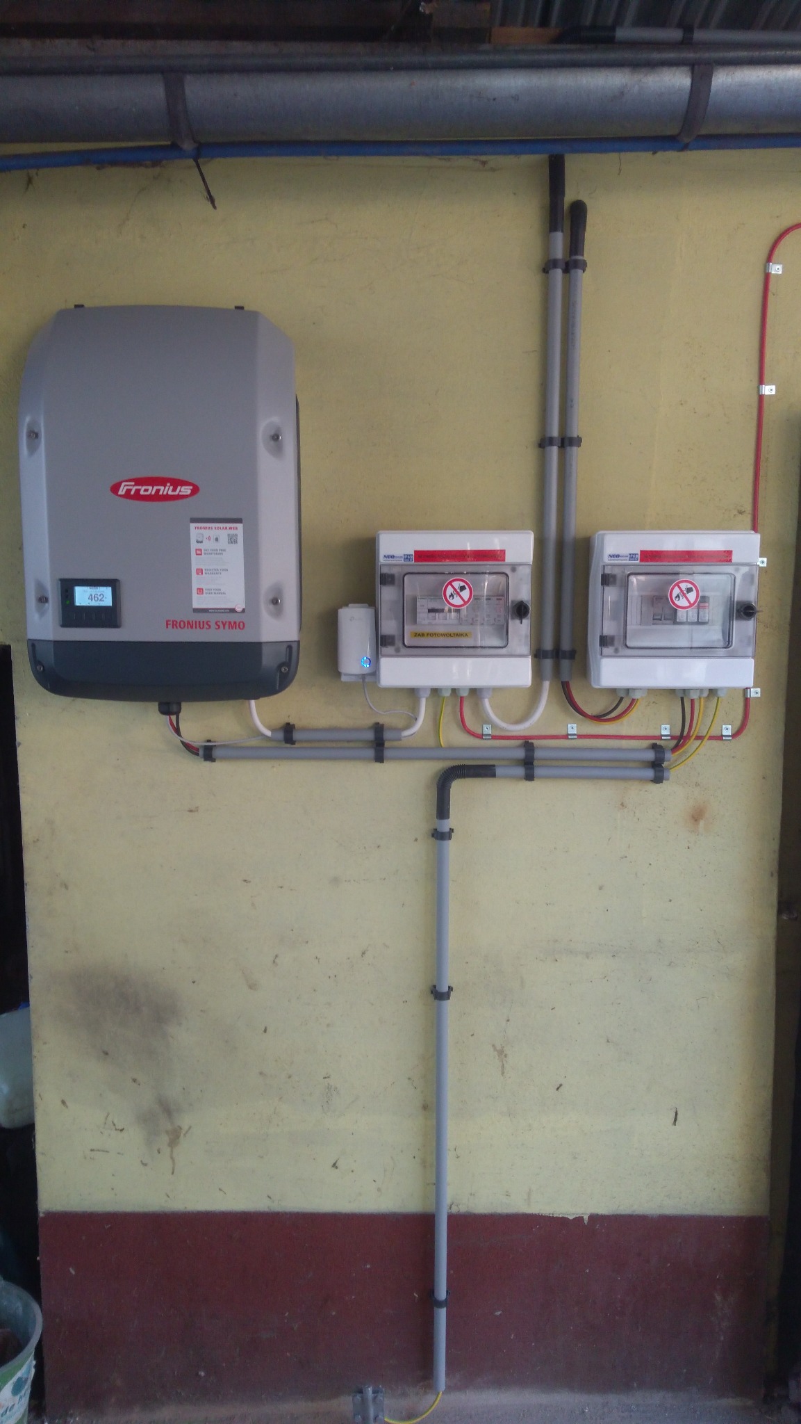 Szary falownik Fronius zamontowany na żółtej ścianie obok dwóch skrzynek zabezpieczeń z naklejkami ostrzegawczymi, połączonych szarymi rurkami PCV z czarnymi uchwytami.