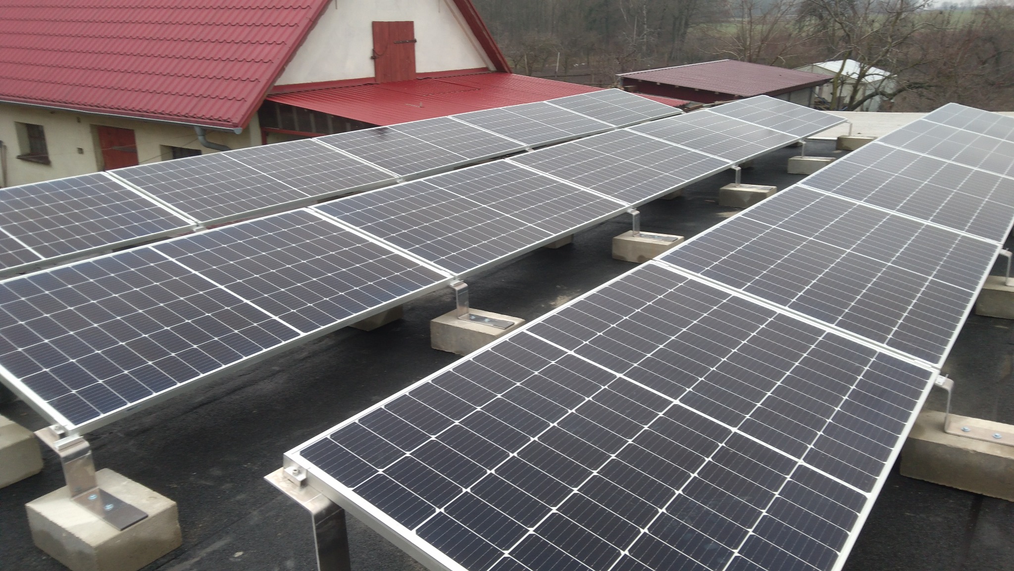 Rząd paneli słonecznych na płaskim dachu, zamontowanych na betonowych podstawach z metalowymi wspornikami, z widocznym budynkiem z czerwoną dachówką w tle.