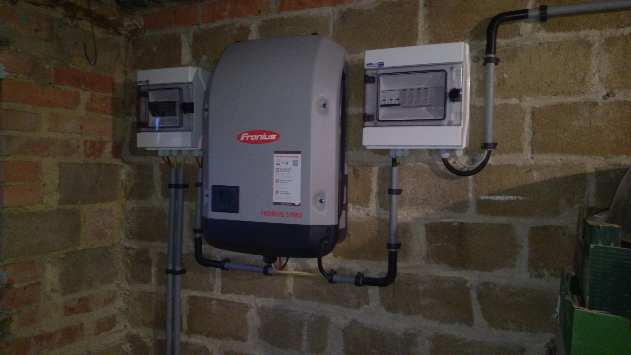 Szary inwerter Fronius Symo zamontowany na ceglanej ścianie piwnicy, obok dwie białe skrzynki rozdzielcze z widocznymi przewodami i rurami instalacyjnymi.