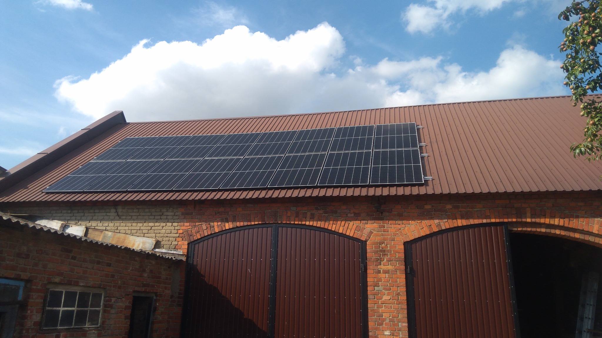 Panele słoneczne zamontowane na brązowym, metalowym dachu budynku z czerwonej cegły z dwoma dużymi, brązowymi bramami garażowymi. Częściowo zachmurzone, błękitne niebo w tle.