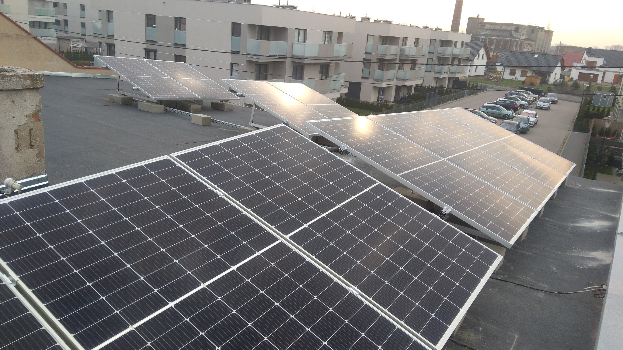 Panele słoneczne zamontowane na płaskim dachu budynku mieszkalnego, widoczne betonowe podstawy i metalowe konstrukcje mocujące, w tle zabudowa osiedlowa i komin przemysłowy.