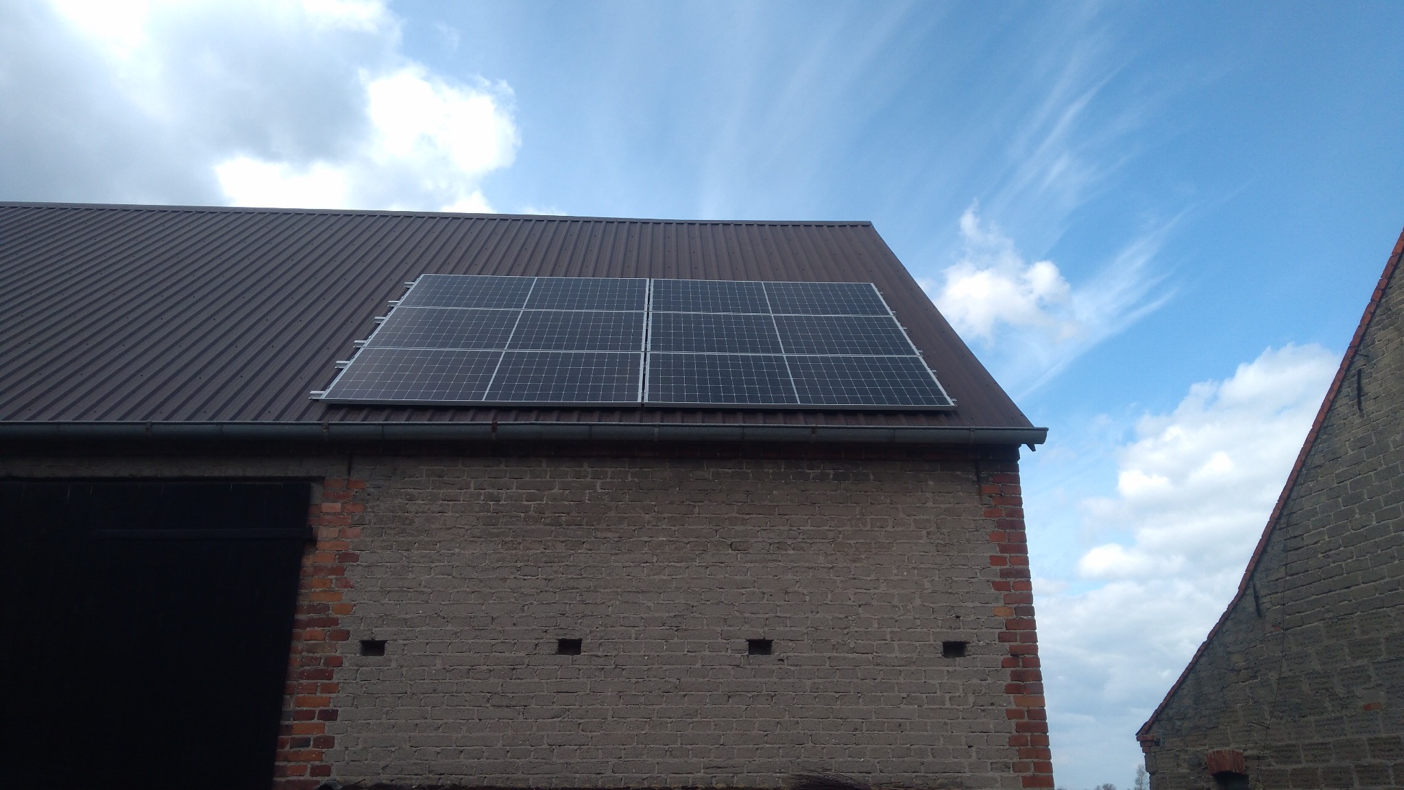 Sześć paneli fotowoltaicznych na falistej, brązowej blasze dachu ceglanego budynku gospodarczego, widok z dołu na błękitne niebo z chmurami.