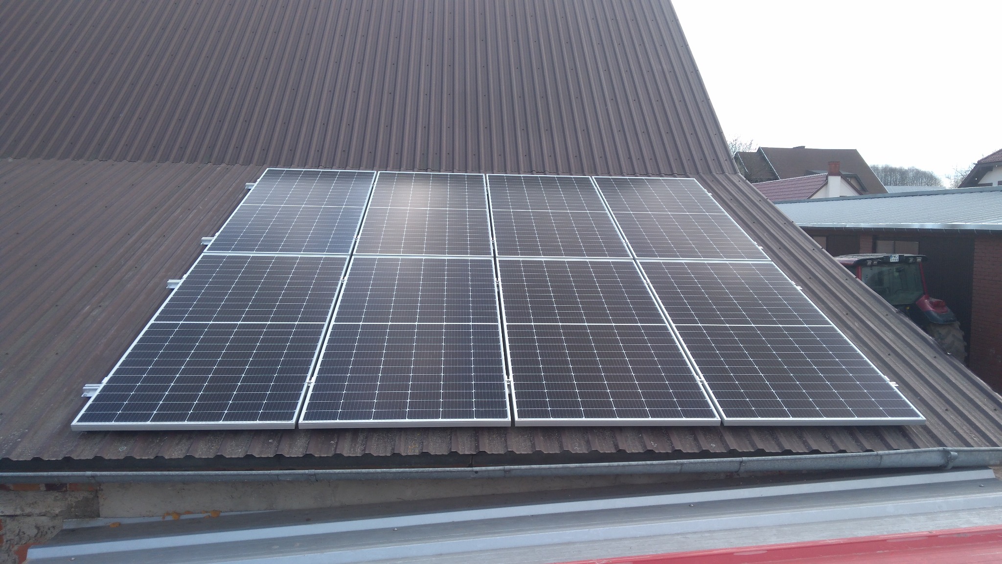 Panel fotowoltaiczny zamontowany na metalowym, falistym dachu budynku gospodarczego, w tle widoczny traktor.