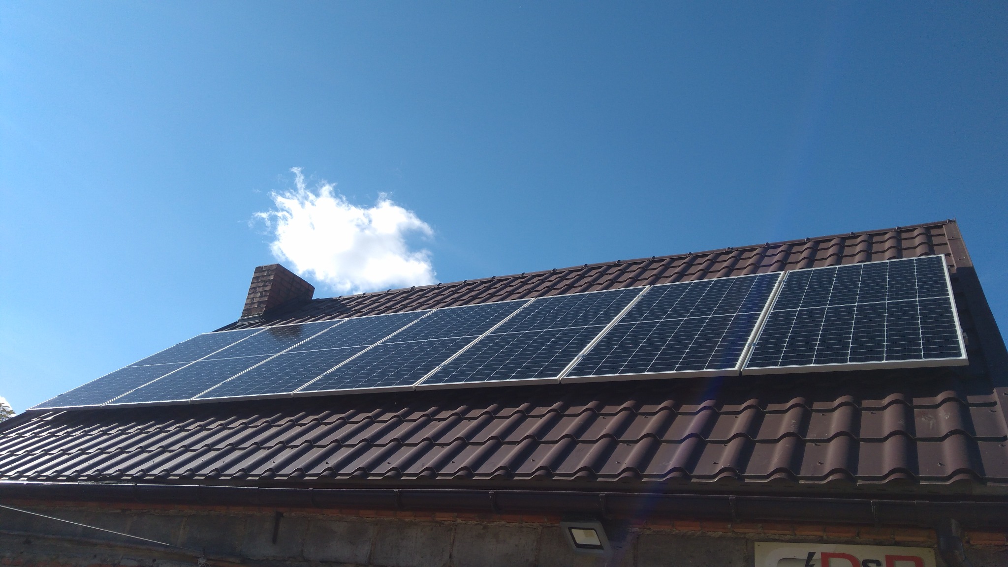 Panele słoneczne zamontowane na brązowym dachu z dachówki ceramicznej, widok z dołu na tle błękitnego nieba z pojedynczą chmurą.