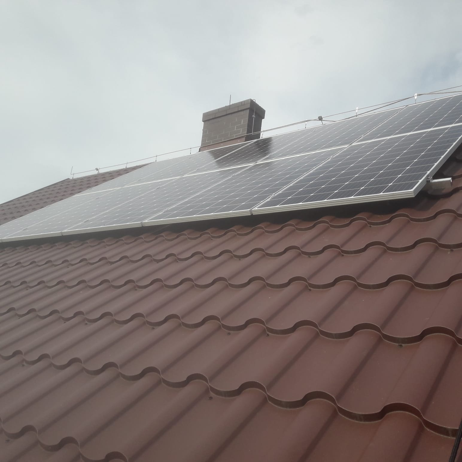 Brązowy dach z dachówki ceramicznej z zamontowanymi panelami fotowoltaicznymi, widoczny komin z cegły i pochmurne niebo.