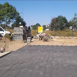 Częściowo ułożona, szara nawierzchnia z kwadratowych elementów, z widocznym stosem materiałów budowlanych, piasku i białym busem w tle, na placu budowy w słoneczny dzień.