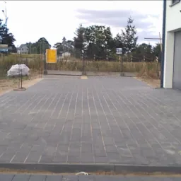 Nowa, szara nawierzchnia z prostokątnych elementów przy budynku z szarą bramą garażową, widoczne palety, samochód i elementy konstrukcyjne w tle.