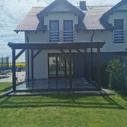 Dwukondygnacyjny dom z ciemną dachówką i nowo wybudowaną drewnianą pergolą na tarasie z szarej kostki, z widoczną drabiną i trampoliną w tle.