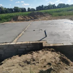 Wylana płyta fundamentowa z wystającą rurą kanalizacyjną, otoczona ceglanym fundamentem, z hałdą piasku w tle i polem kukurydzy w oddali.