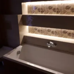 Biała wanna z chromowaną baterią, podświetlane półki z dekoracyjną tapetą w liście, toaleta w tle, ciemne oświetlenie