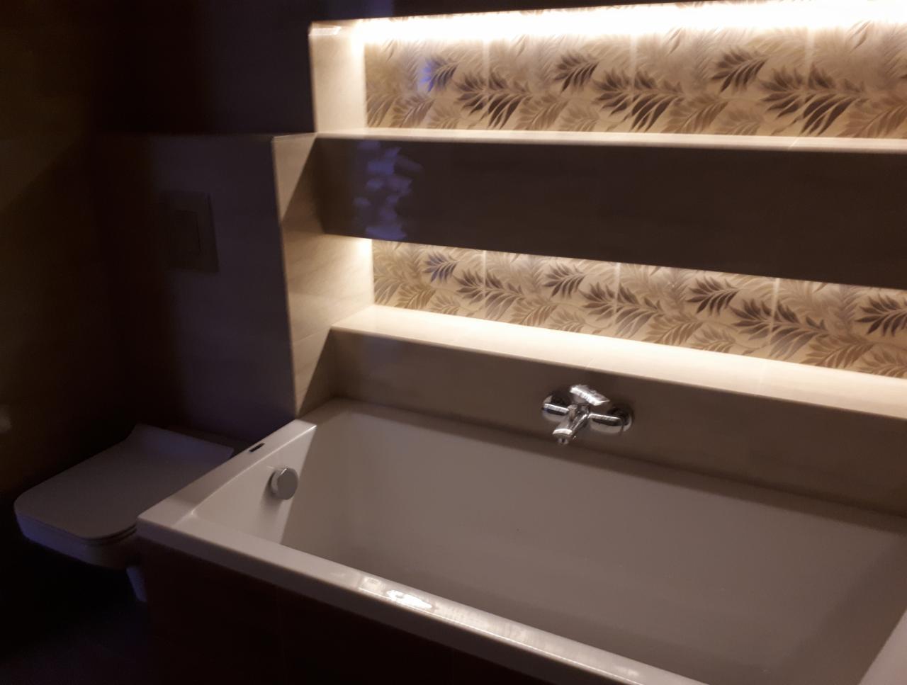 Biała wanna z chromowaną baterią, podświetlane półki z dekoracyjną tapetą w liście, toaleta w tle, ciemne oświetlenie