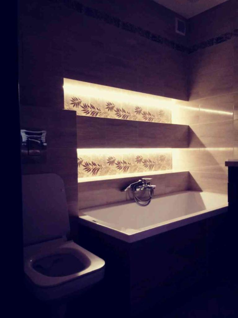 Nowoczesna łazienka z podświetlanymi półkami, beżowe płytki z motywem roślinnym, biała wanna i toaleta w odcieniach szarości, ciemne oświetlenie.