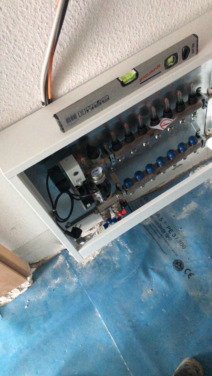 Rozdzielacz instalacji grzewczej w białej obudowie, poziomowany poziomicą PROJAHN, widoczne przewody elektryczne i fragment niebieskiej folii izolacyjnej na podłodze.