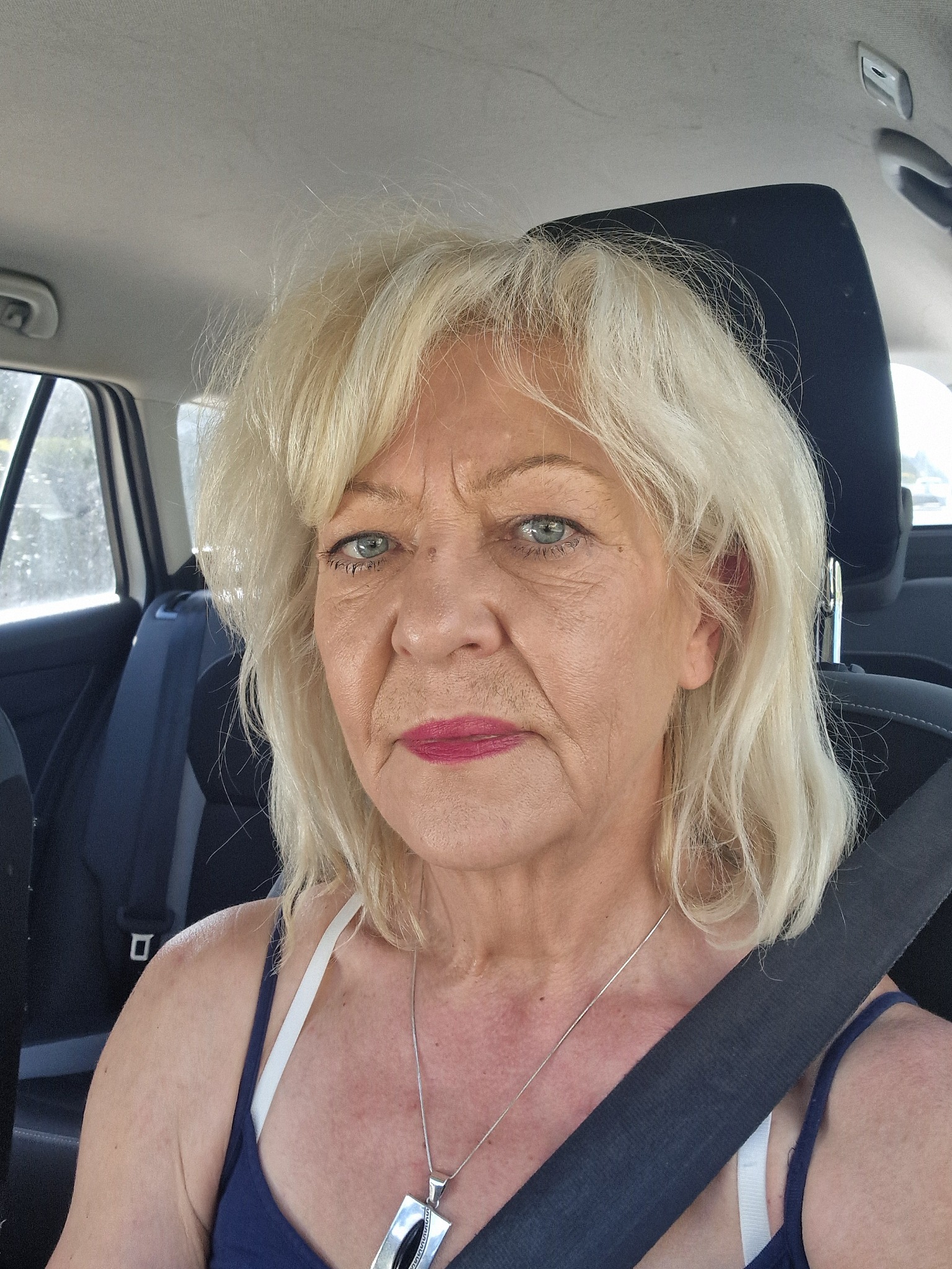 Portret kobiety z blond włosami, siedzącej w samochodzie z zapiętym pasem bezpieczeństwa i srebrnym naszyjnikiem.