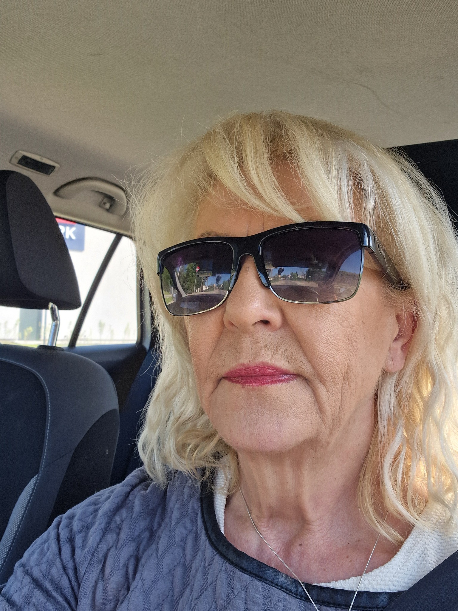 Portret kobiety w okularach przeciwsłonecznych, siedzącej w samochodzie; w odbiciu okularów widoczny budynek i drzewa.