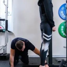 Asystent asekuruje kobietę podczas ćwiczenia na siłowni, zwracając uwagę na ustawienie stóp i kolan.