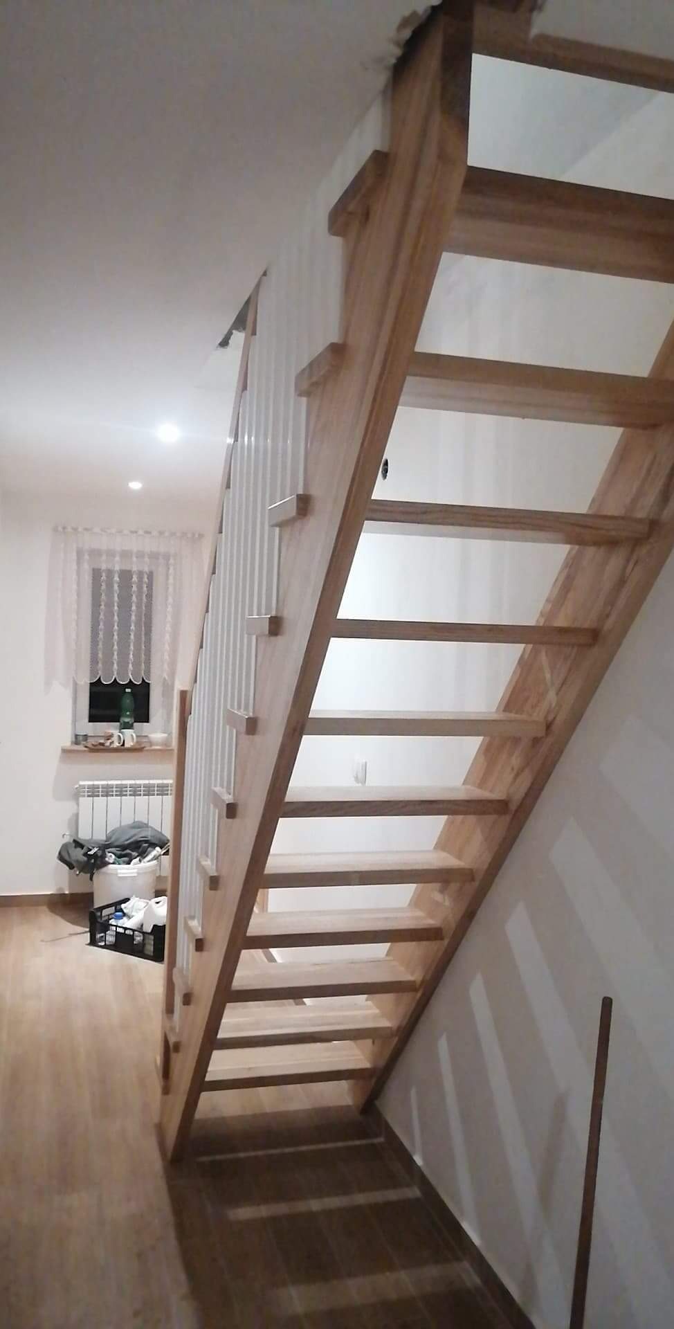 Jasne, drewniane schody z białymi balustradami w trakcie montażu, widoczne niedoskonałości wykończenia sufitu i ściany.