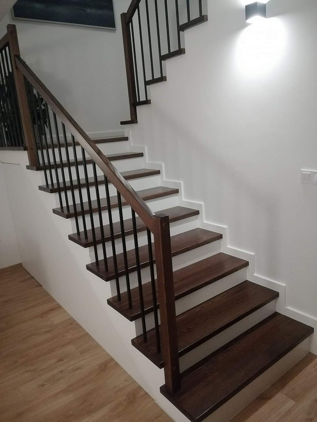 Nowoczesne schody z ciemnego drewna z czarnymi metalowymi balustradami, białe podstopnice i listwy przypodłogowe, oświetlenie punktowe na ścianie.