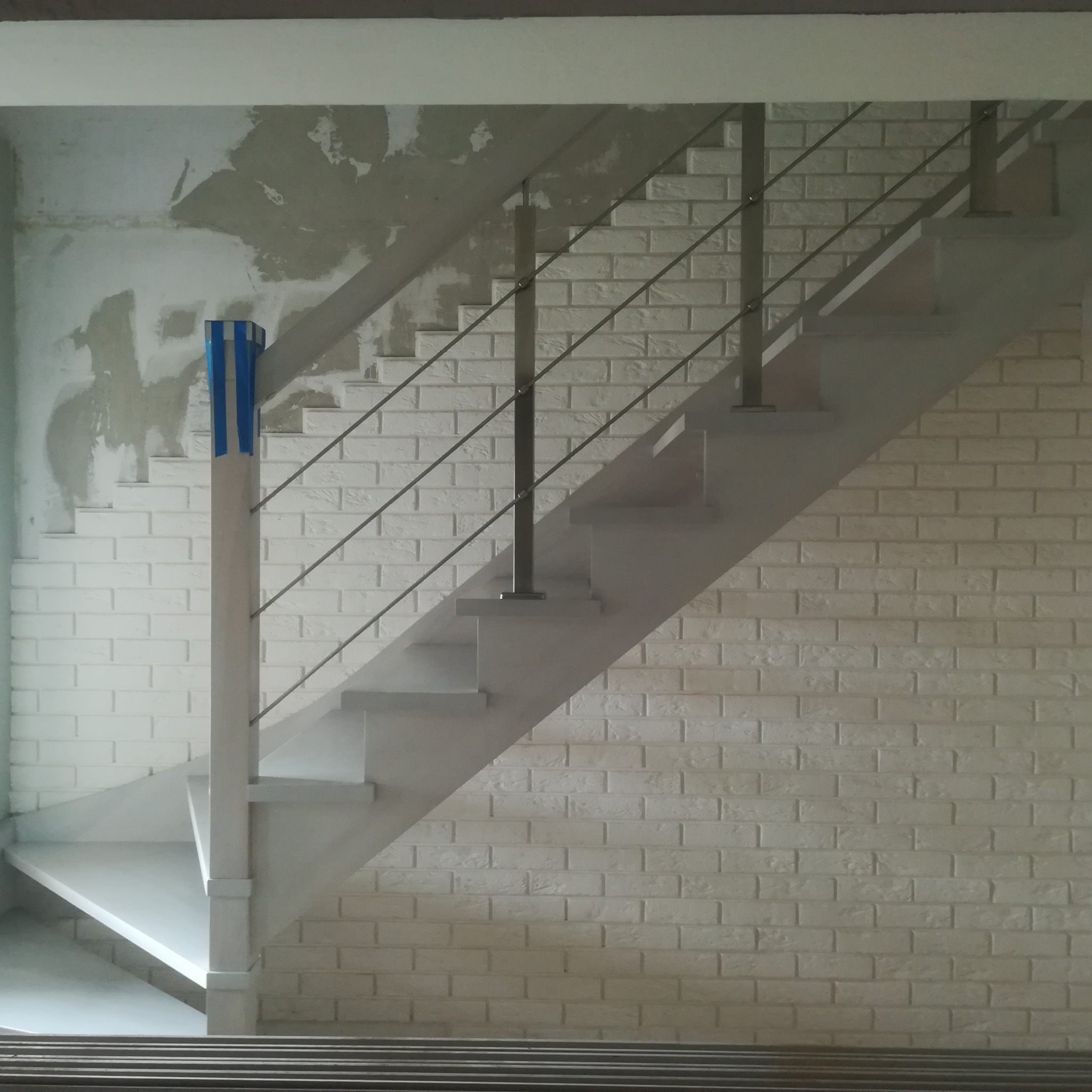 Betonowe schody ze stalową balustradą, zamontowane przy ścianie wyłożonej cegłami, z drewnianym słupkiem zabezpieczonym niebieską taśmą malarską.