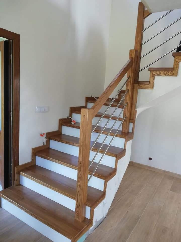 Jasne, drewniane schody z białymi podstopnicami i metalową balustradą, widok z dołu na tle jasnych ścian i drewnianej podłogi.