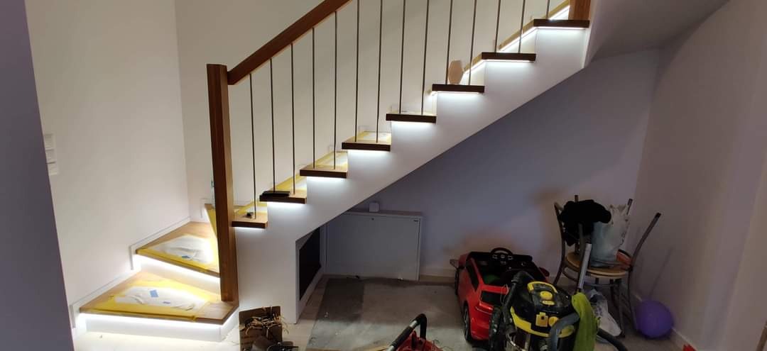 Drewniane schody z podświetleniem LED na stopniach, balustrada z drewnianą poręczą i czarnymi prętami, widoczne narzędzia i zabawki w przestrzeni pod schodami.