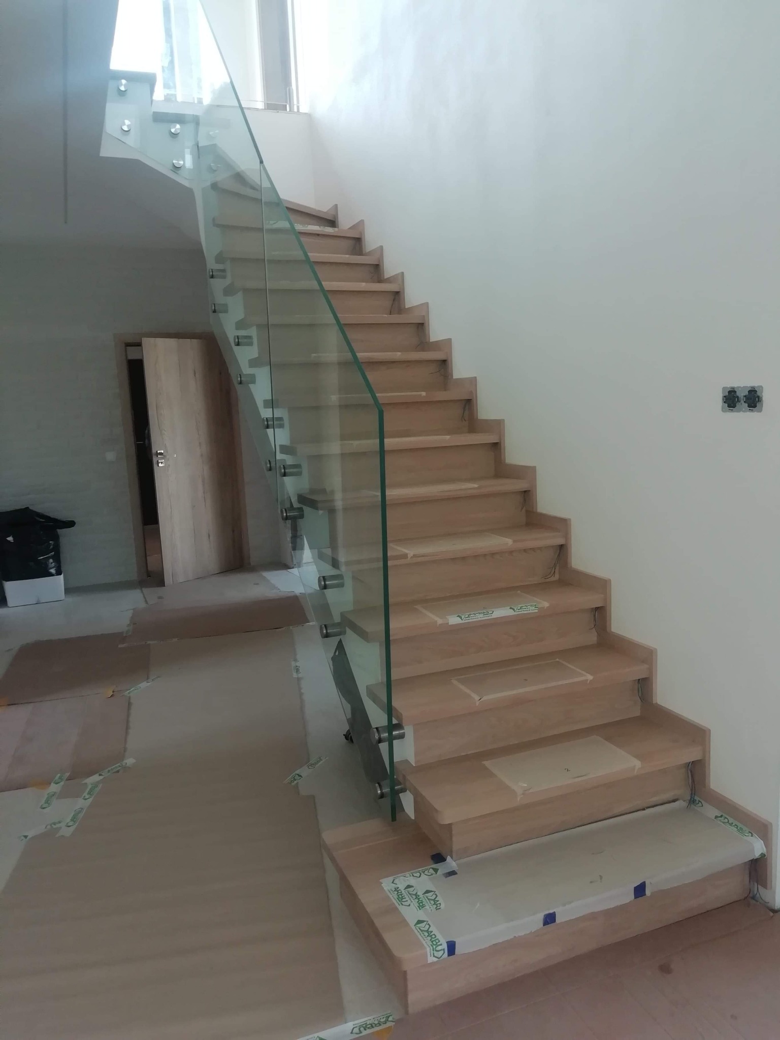 Jasne, drewniane stopnie schodów z przezroczystą szklaną balustradą, zabezpieczone folią ochronną, w trakcie montażu.