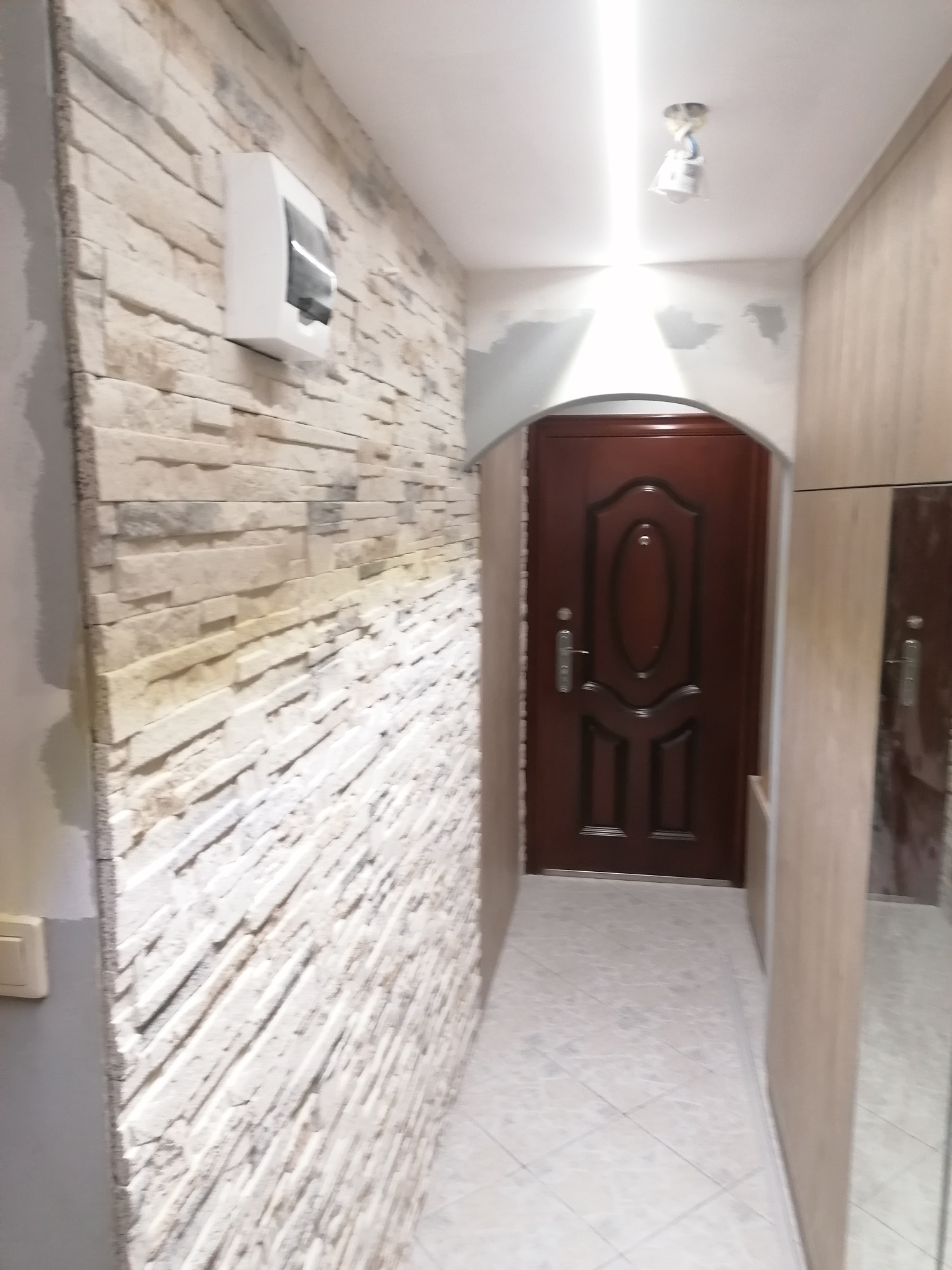 Korytarz po remoncie: ściana wyłożona dekoracyjnym kamieniem w odcieniach beżu, brązowe drzwi wejściowe z ozdobnym wzorem i biała skrzynka elektryczna na ścianie.