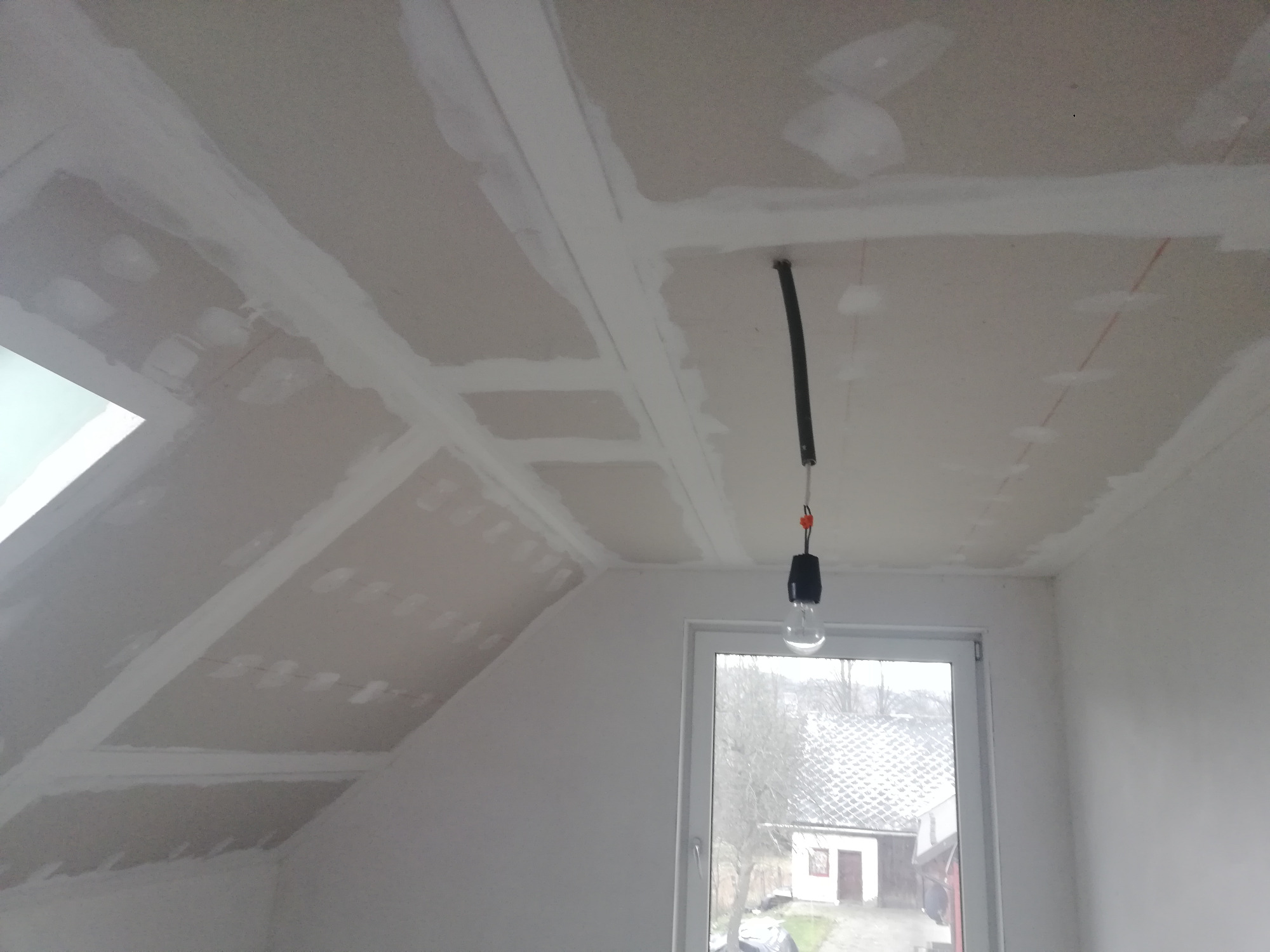 Poddasze w trakcie wykańczania: zamontowane płyty gipsowo-kartonowe na skosach i suficie, widoczne łączenia płyt zaszpachlowane, wisząca żarówka na kablu, okno z widokiem na zewnątrz.