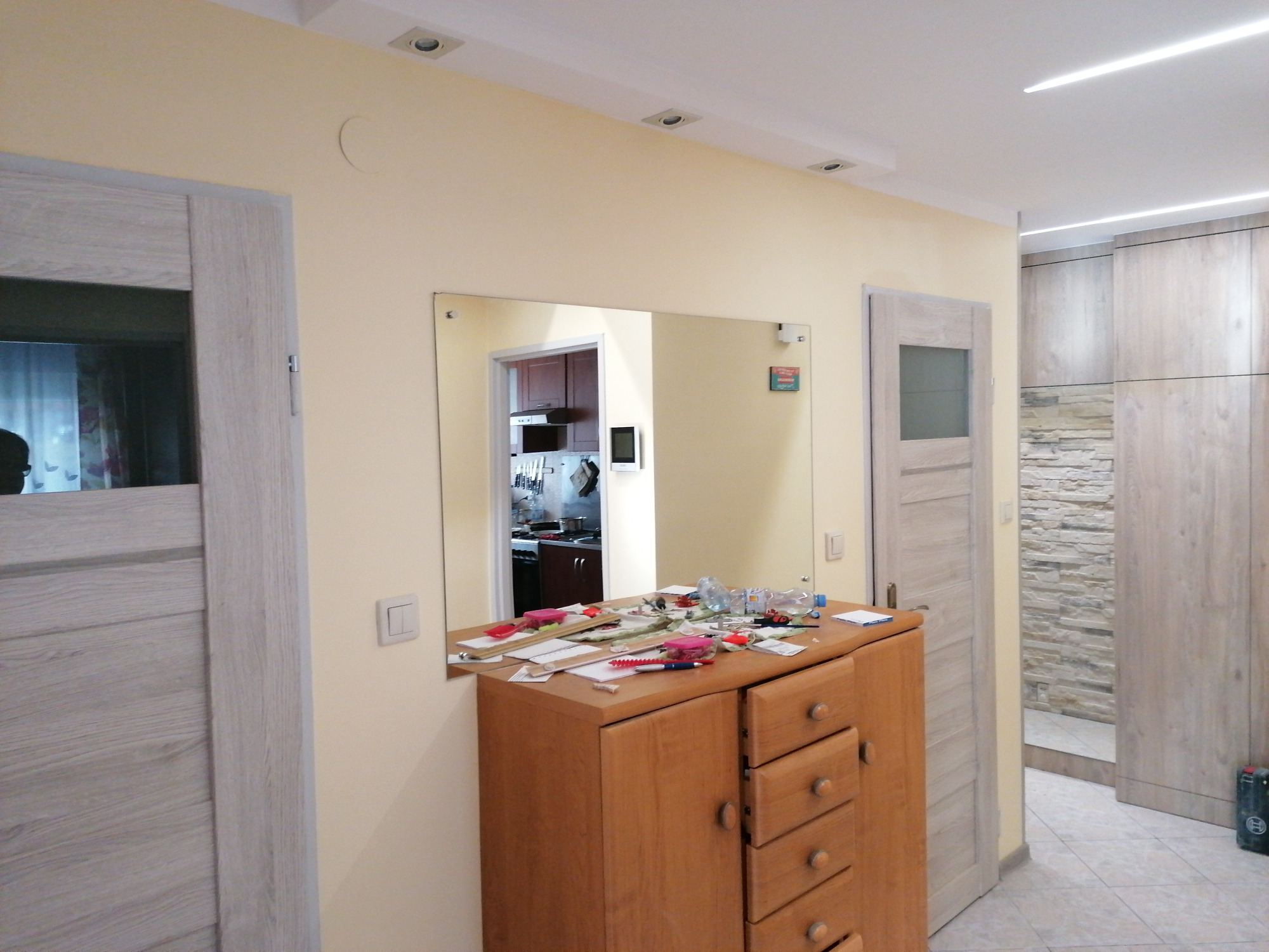 Umeblowane wnętrze mieszkania z widoczną szafką z lustrem, na której leżą różne przedmioty, oraz fragmentem kuchni w odbiciu lustrzanym.