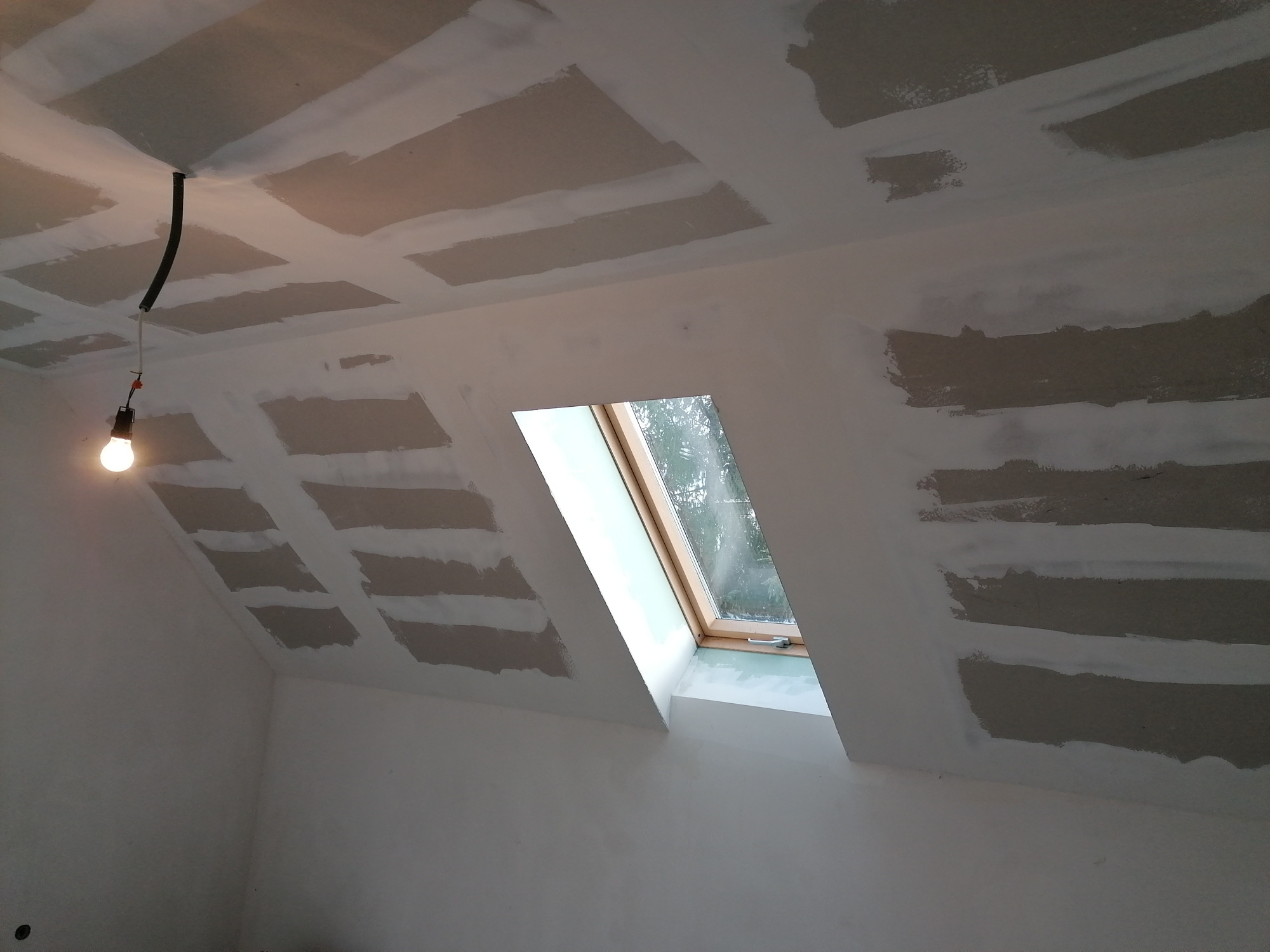Poddasze w trakcie wykańczania: widoczne płyty gipsowo-kartonowe na skosach sufitu, łączenia szpachlowane, okno dachowe, wisząca żarówka na kablu.