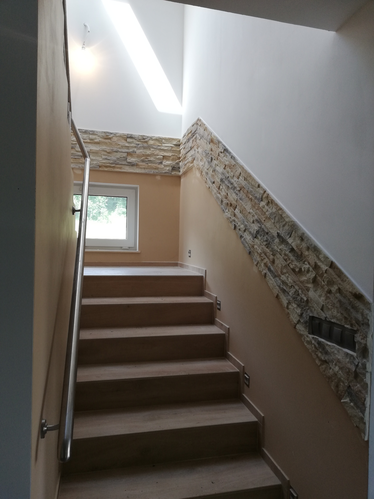 Klatka schodowa w Sucha Beskidzka z drewnianymi schodami i dekoracyjną ścianą z kamienia, oświetlona naturalnym światłem z okna i świetlika.