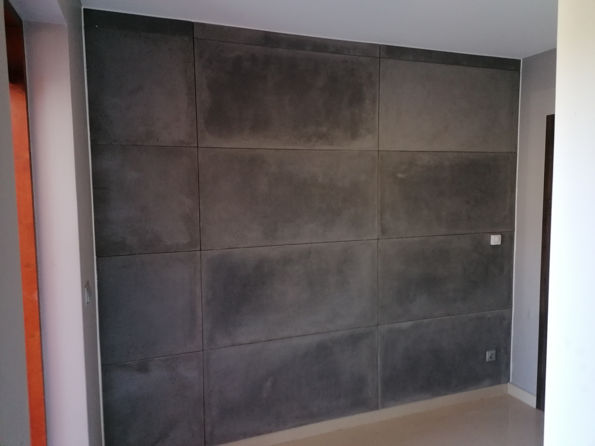 Ściana w pomieszczeniu wykończona panelami imitującymi beton architektoniczny, ułożonymi w regularną siatkę, widoczny włącznik światła i gniazdko elektryczne.