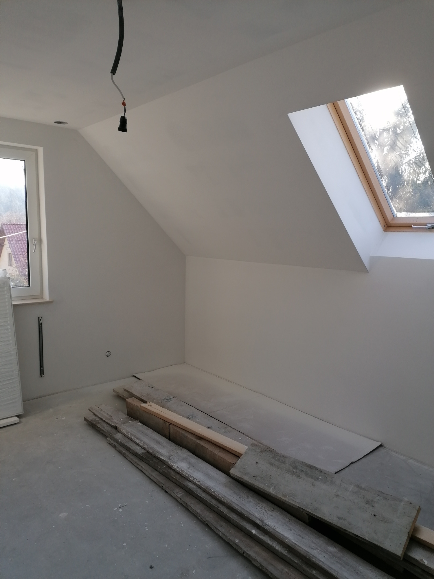 Poddasze po gładziowaniu ścian, przygotowane do dalszych prac wykończeniowych; widoczne drewniane deski na podłodze, okno dachowe z drewnianą ramą i surowa instalacja elektryczna.