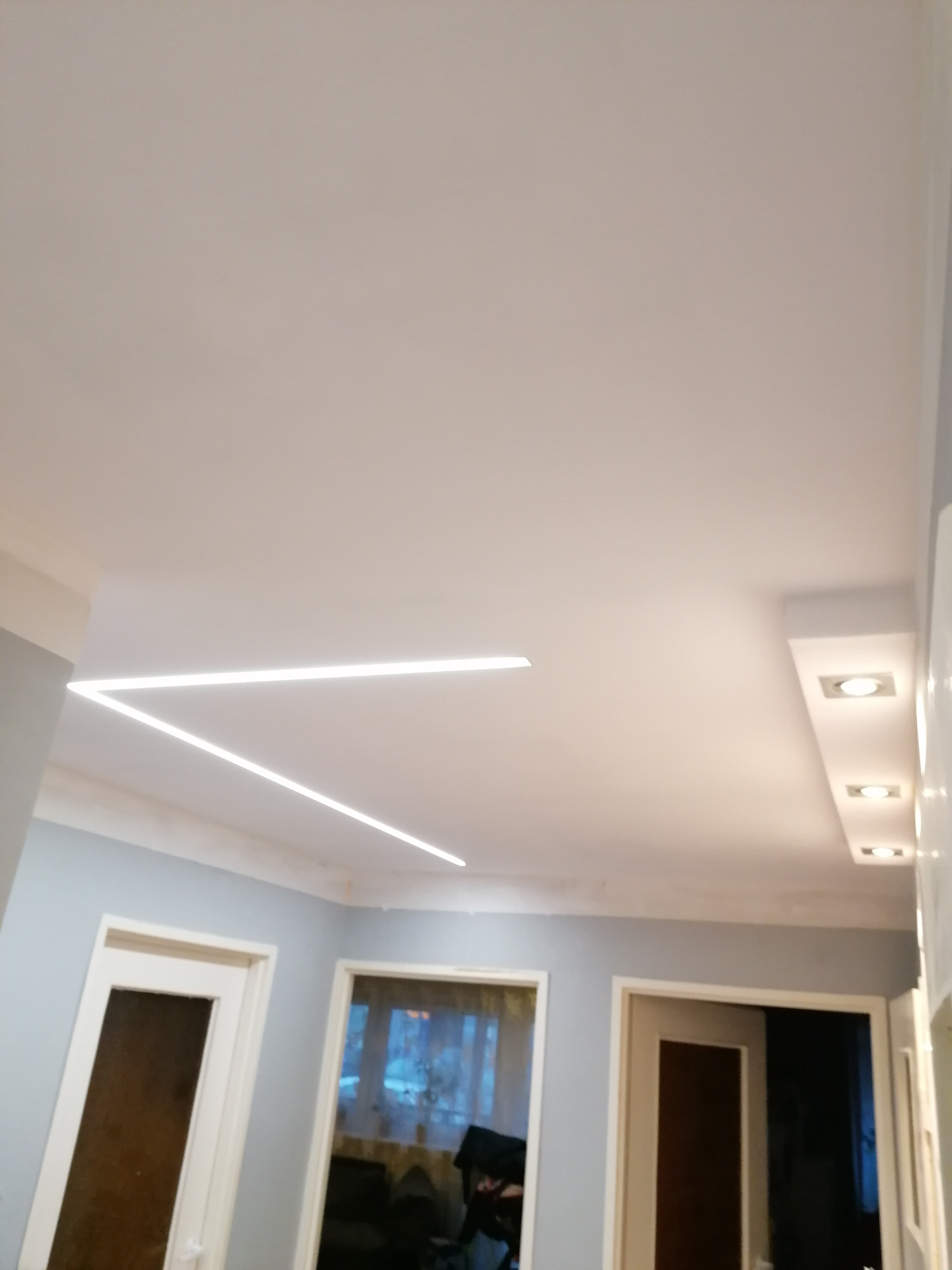 Nowoczesne oświetlenie sufitowe w kształcie litery V, z dodatkowymi punktowymi lampami w zabudowie, szare ściany i białe futryny drzwi, widok z dołu.