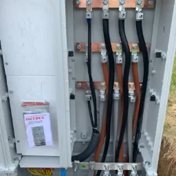 Wnętrze skrzynki elektrycznej z zamontowanymi przewodami o różnych kolorach (czarny, brązowy, niebieski, żółto-zielony) podłączonymi do miedzianych szyn zbiorczych, widoczna dokumentacja techniczna...