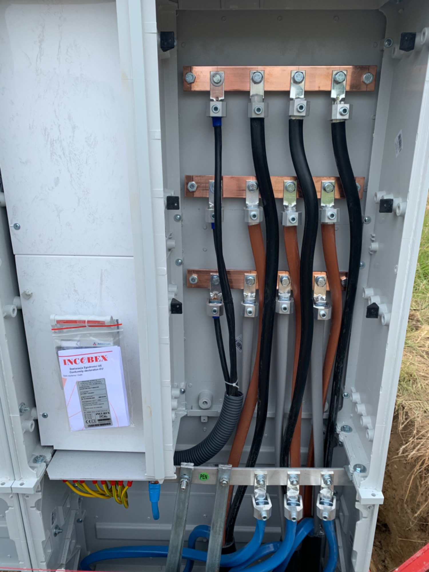 Wnętrze skrzynki elektrycznej z zamontowanymi przewodami o różnych kolorach (czarny, brązowy, niebieski, żółto-zielony) podłączonymi do miedzianych szyn zbiorczych, widoczna dokumentacja techniczna...