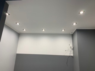 Biały sufit z równomiernie rozmieszczonymi, kwadratowymi oprawami oświetleniowymi LED, szara ściana w dolnej części, widoczne przewody elektryczne.