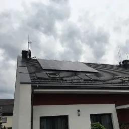 Panele fotowoltaiczne na dachu nowoczesnego domu jednorodzinnego z ciemną dachówką i oknami dachowymi, pochmurne niebo w tle.