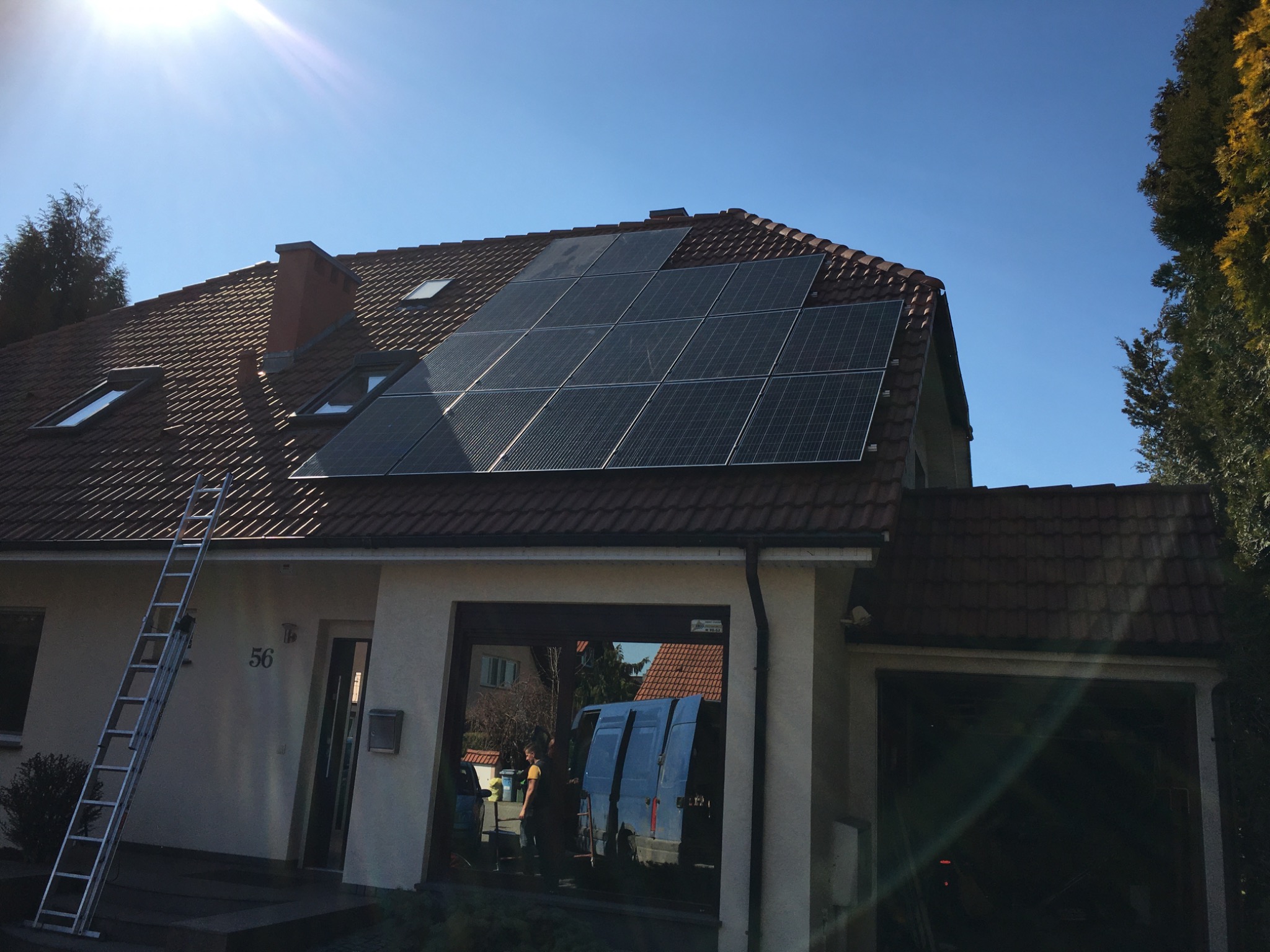 Dom jednorodzinny z zamontowanymi panelami słonecznymi na dachu z czerwonej dachówki, drabina oparta o ścianę, widoczny samochód dostawczy w garażu.