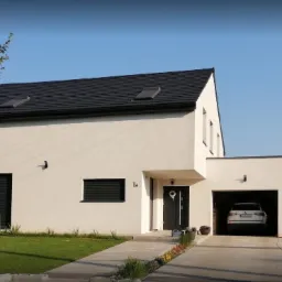 Nowoczesny dom murowany z garażem w bryle budynku, czarny dach z oknami dachowymi, jasna elewacja, zadbany trawnik i podjazd z kostki.